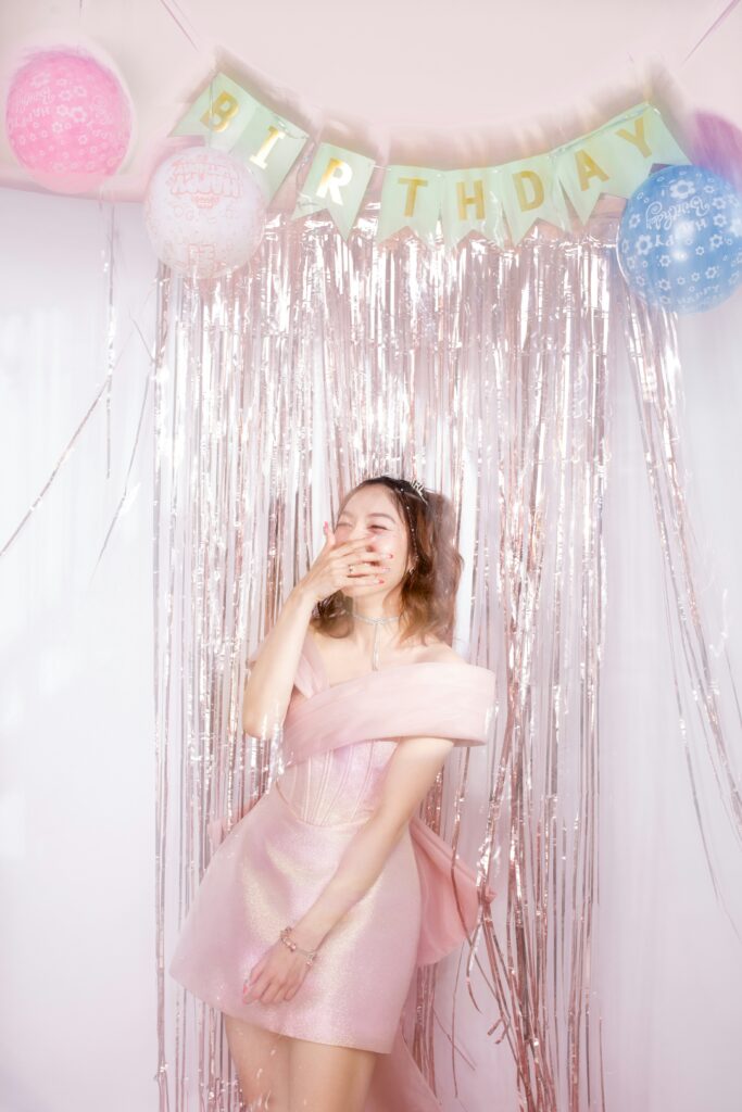 young woman wearing a pink dress, laughing in front of pink birthday decorations.